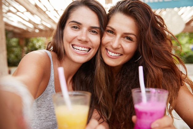 Cheerful positive women pose at camera of unrecognizable device, make selfie as drinks fresh colourful cocktails, sits against outdoor cafe interior.