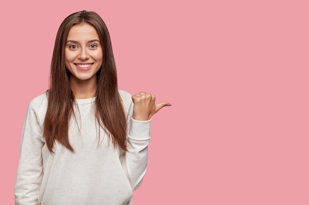 Free photo cheerful positive young european woman with dark hair, broad shining smile, points with thumb aside