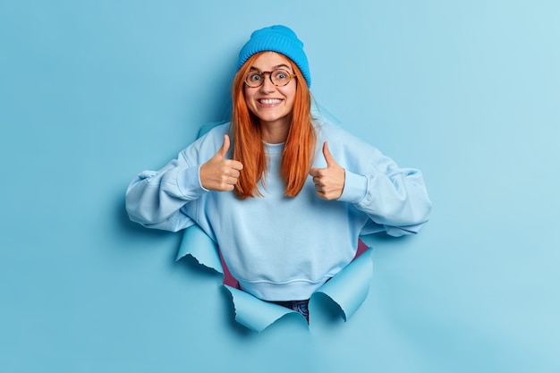 Free photo cheerful redhead european woman makes thumb up gesture makes excellent sign approves something smiles broadly wears hat and sweater breaks through paper hole