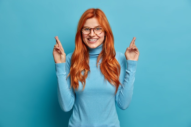 Cheerful redhead European woman smiles gladfully crosses fingers makes wish hopes to receieve positive results dressed in casual turtleneck.