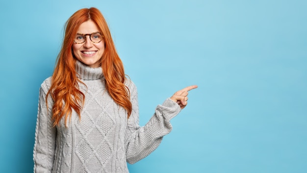 cheerful redhead European woman in spectacles smiles broadly and points away on copy space gives direction to best shop wears warm winter sweater.