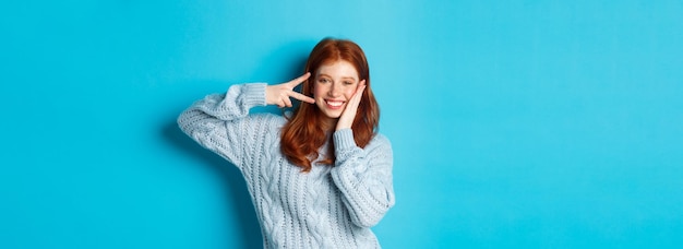 Free photo cheerful redhead female model sending good vibes smiling and showing peace sign standing over blue b