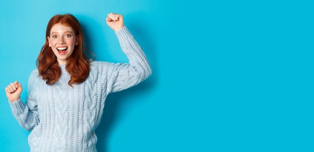 Free photo cheerful redhead gil winning celebrating victory smiling and jumping from happiness posing against b