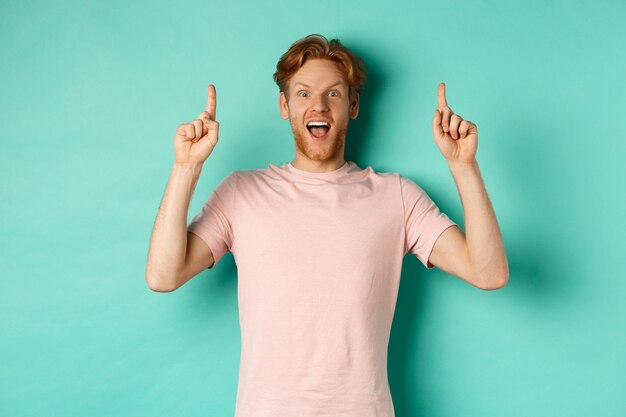 Cheerful redhead man in t-shirt pointing fingers up, staring in awe at camera and showing advertisement, standing over turquoise background.