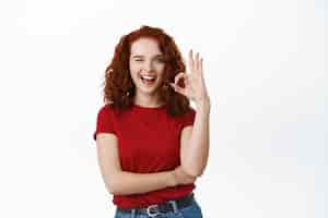 Free photo cheerful redhead woman with curly hair showing ok sign, winking and smiling, say yes, encourage to buy something, make alright gesture, approve and praise good quality product