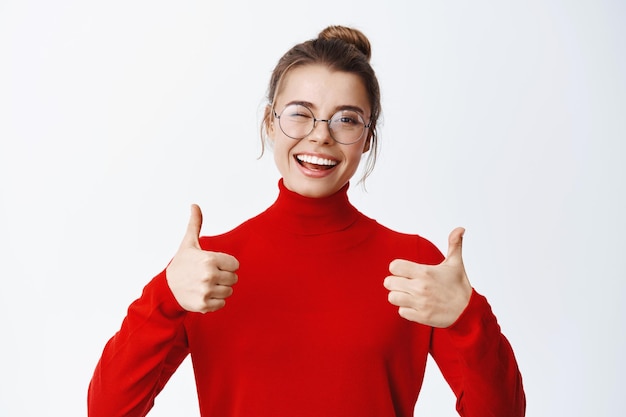 Free Photo cheerful smiling female student in glasses showing thumbs up in approval, praise and recommend good thing, approve and like it, standing on white