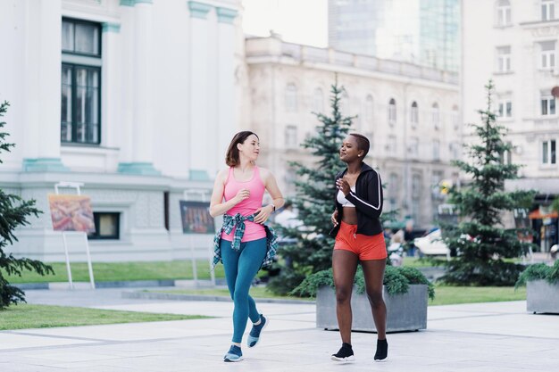 Cheerful smiling friends in sportswear running in the city dicussing Multiethnic women having a fitness workout