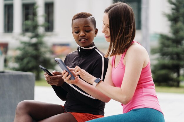 Cheerful smiling friends in sportswear sitting on a bench in the city discussing while using smartphone looking to screen Multiethnic women having a fitness workout break