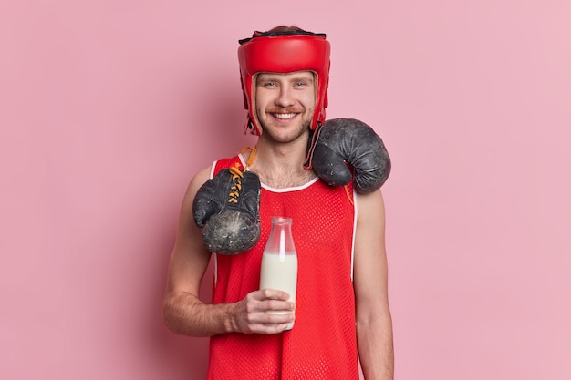 Free Photo cheerful sportsman with mustache and happy smile on face wears protective hat carries boxer gloves around neck drinks milk from glass bottle for having muscles builds body and character.