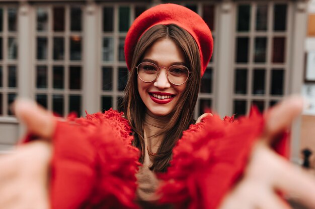 Cheerful straighthaired browneyed girl in stylish glasses wearing red beret and woolen jacket wants to hug