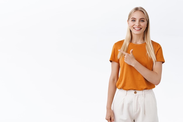 Free Photo cheerful stylish blond caucasian woman in orange tshirt smiling enthusiastic pointing finger left at blank white space giving advice what choose standing white background joyful