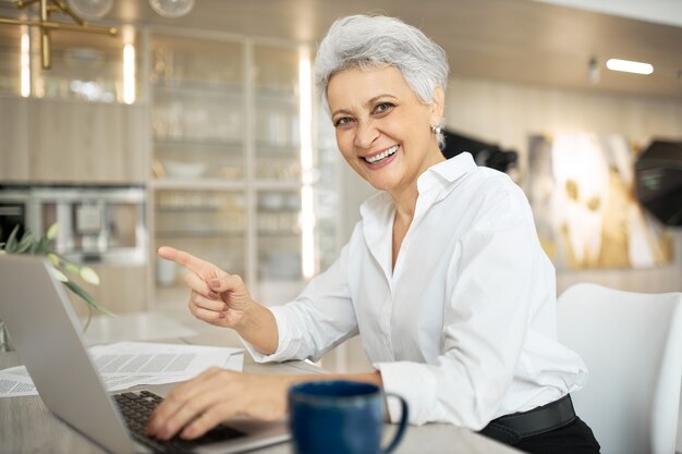 Cheerful stylish mature female manager working in office, sitting at desk with generic portable computer, smiling broadly, pointing fore finger, enjoying her job