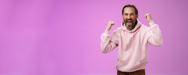 Free photo cheerful supportive manly mature adult bearded guy fan yelling raising clenched fists triumphing team scored goal celebrating standing pleased shouting achieving success posing purple background