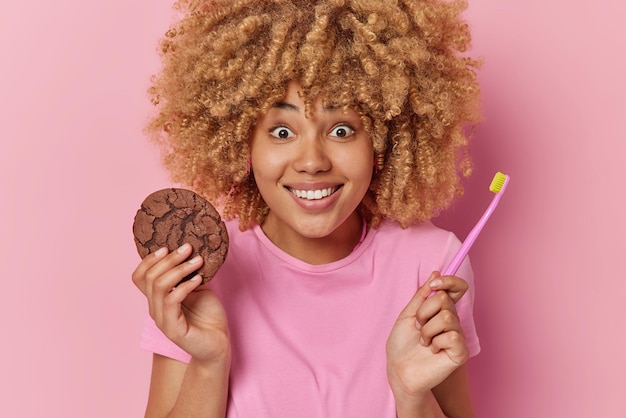 Cheerful surprised curly haired woman holds delicious chocolate cookie eats harmful food with bad influence on teeth poses with toothbrush wears casual t shirt isolated over pink background