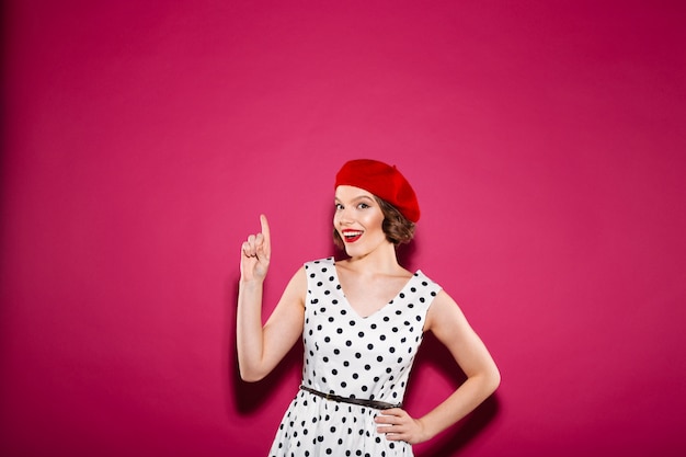 Cheerful surprised ginger woman in dress with arm on hip pointing up and looking at the camera over pink