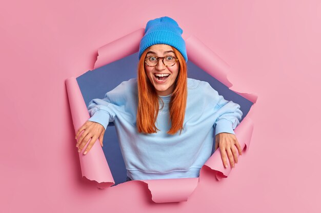 Cheerful surprised redhead woman looks through torn paper, through ripped hole.