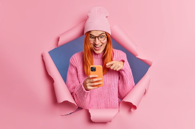 Cheerful surprised redhead woman points at smartphone screen reacts on big discounts in online store laughs happily and looks at something unbelievable wears stylish clothes.