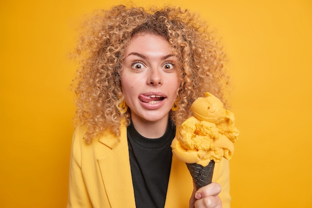 Free photo cheerful surprised woman with curly bushy hair licks lips sticks out tongue holds big delicious ice cream tempting to eat summer dessert poses against vivid yellow wall. mmm how tasty.