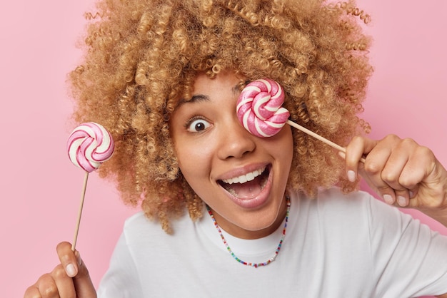 Free photo cheerful surprised woman with curly hair holds two caramel candies on sticks enjoys eating sweet snack smiles broadly wears casual white t shirt poses against pink background taste these lollipops