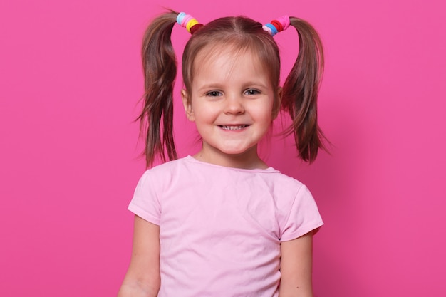 Free Photo cheerful sweet little girl with funny pigtails, smiling sincerely, standing straight, having colourful scrunchies. copy space for advertisement.