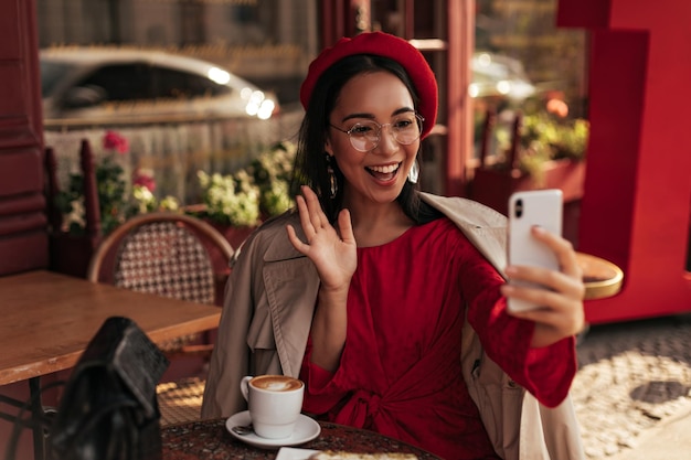 Free Photo cheerful tanned brunette asian woman in beige trench coat red dress and beret smiles waves hand in greeting and talks by video in her cellphone charming lady sits in street cafe