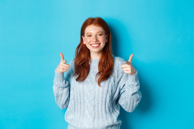 Free photo cheerful teen girl with red hair, pointing fingers at camera and smiling, congratulating or praising you, standing over blue background