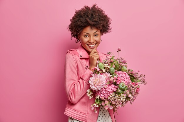 Cheerful tender young Afro American woman being in high spirit, gets pretty flowers, enjoys nice gift, smiles broadly, poses indoor. Happy teacher receives bouquet from pupils on Day of Knowledge