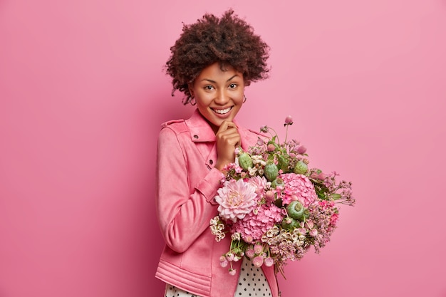 Cheerful tender young Afro American woman being in high spirit, gets pretty flowers, enjoys nice gift, smiles broadly, poses indoor. Happy teacher receives bouquet from pupils on Day of Knowledge