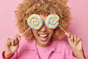 Free photo cheerful unrecognizable curly woman covers eyes with sweet lollipops on sticks has sugar addiction enjoys cheat meal day dressed in casual clothes poses against pink background harmful food