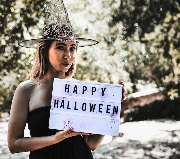 Cheerful witch holding sigboard with greeting in sunny woodland