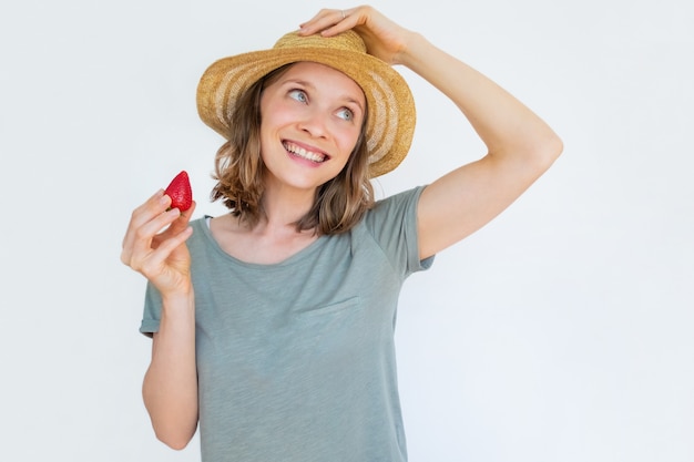 Cheerful woman in hat holding ripe strawberry and smiling