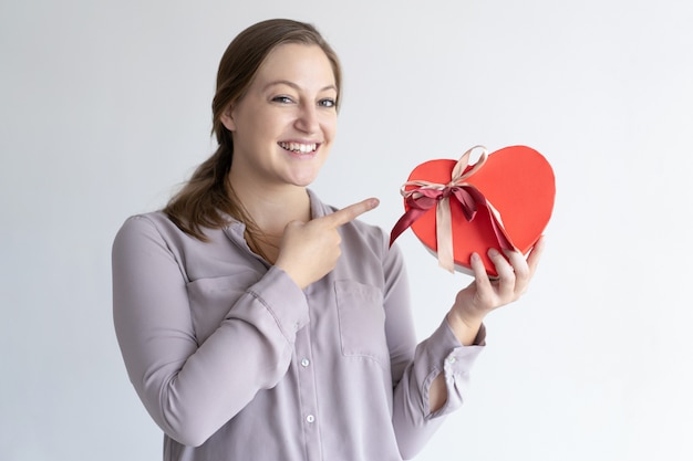 Free photo cheerful woman holding heart shaped gift box and pointing at it