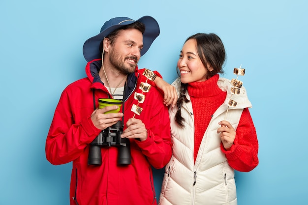 Free photo cheerful woman and man have camping trip together, hold tasty marshmallow made on bonfire, look with smile at each other, spend leisure time in wild nature carry binoculars