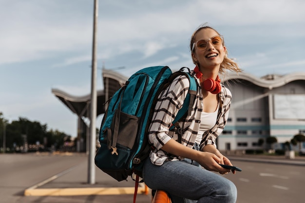 Free photo cheerful woman in plaid shirt and denim pants smiles widely near airport blonde girl in sunglasses holds backpack and phone tourist in denim pants and white tee poses