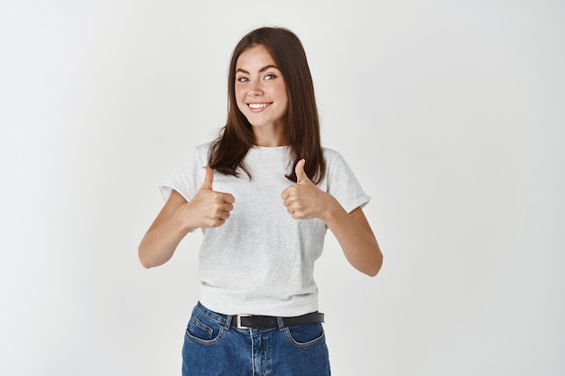 Free Photo cheerful woman saying yes, showing thumbs-up in approval, like and agree with you, standing over white wall.