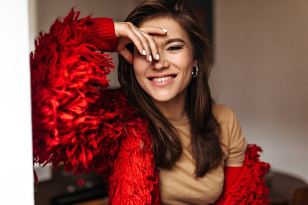 Cheerful woman in silver earrings, knitted coat and beige top laughs, leaning on white wall.