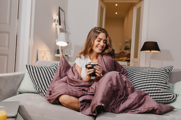 Free photo cheerful woman sitting on couch with blanket and cushions and smiling