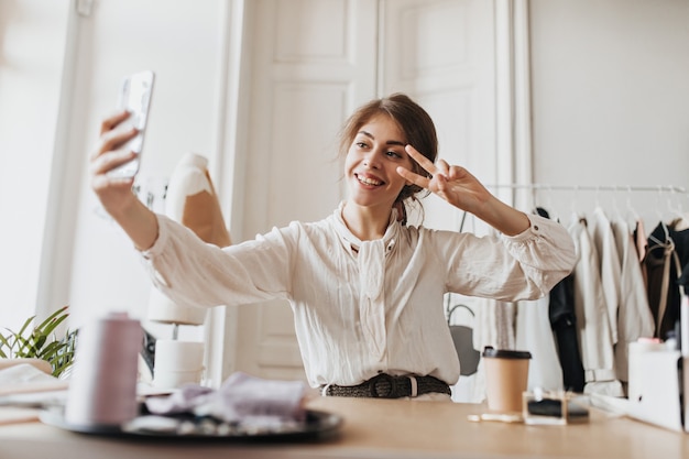 Cheerful woman in stylish blouse taking selfie and showing peace sign