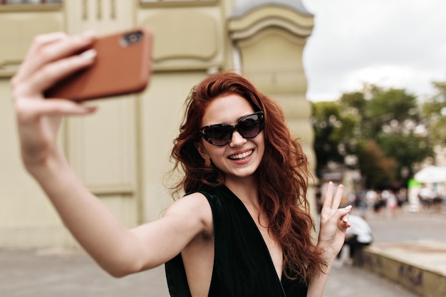 Cheerful woman in sunglasses showing peace sign and taking selfie outside