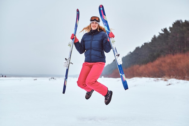 Free photo cheerful woman wearing warming sportswear holding skis and jumping on a snowy beach