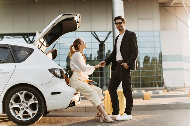 Cheerful woman in white blouse beige pants sits in car and holds boyfriend hand Brunette man in black suit and eyeglasses smiles and poses near airport