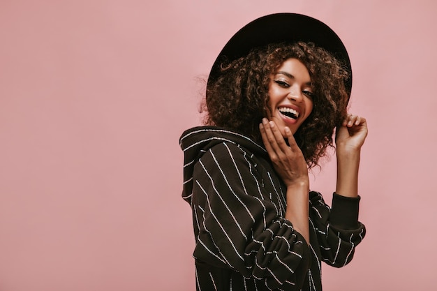 Cheerful woman with dark wavy hairstyle in black striped outfit and hat laughing and looking into camera on pink background