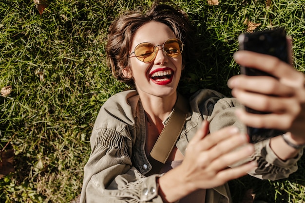 Free photo cheerful woman with short hair in yellow glasses laughs and lies on grass outside. woman in denim jacket making selfie outdoors.