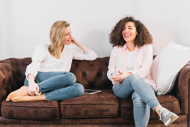 Free photo cheerful women with technologies speaking on sofa