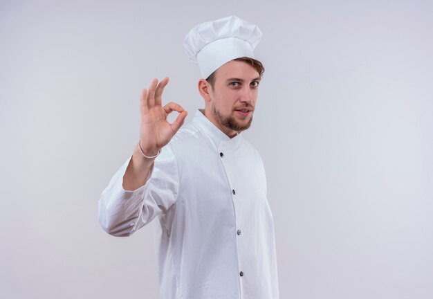 A cheerful young bearded chef man in white uniform showing ok gesture on a white wall