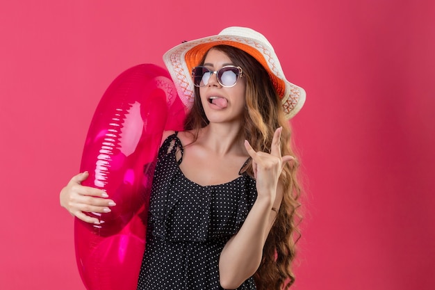 Free photo cheerful young beautiful traveler girl in dress in polka dot in summer hat wearing sunglasses holding inflatable ring happy and positive sticking out tongue making victory sign standing over pi