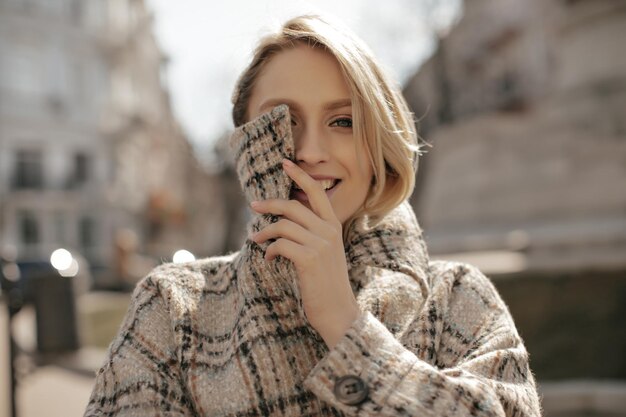 Cheerful young blonde woman smiles sincerely outdoors and covers part of her face with collar of tweed checkered coat