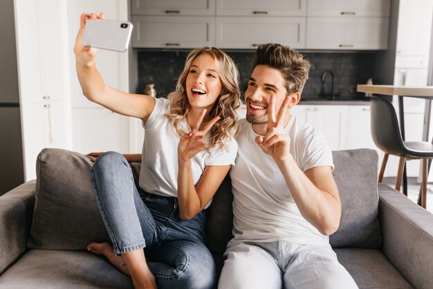 Cheerful young couple at home making selfie with peace sign and smiling widely. Happy girl sitting on the couch with boyfriend and taking portrait of them together.