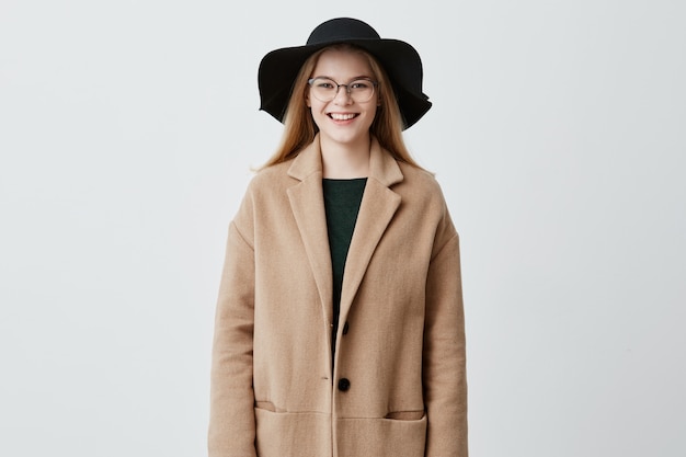 Cheerful young European woman with beautiful set of features, dressed in coat over green sweater, wearing her blonde hair loose, with eyeglasses on, looking  with charming smile