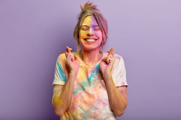 Cheerful young European woman with satisfied facial expression, focuses on wish, crosses fingers, dirty with colorful Holi powder, hopes over purple wall, comes on color festival with friends.